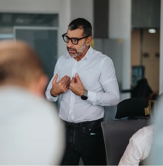 Person in white shirt speaks with commitment in front of colleagues in a modern office; focus on communication, leadership and personal presence in a business context.