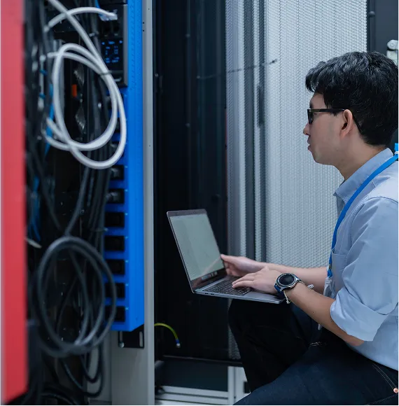 Person works with laptop in the server room next to network hardware; focus on IT infrastructure, maintenance and data security.