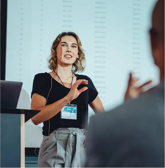 Fachvortrag auf Schulungs-Konferenz Person hält eine Präsentation mit Headset-Mikrofon auf einer Konferenzbühne vor großem Bildschirm; Fokus auf Fachvortrag, professionelles Auftreten und Expertenwissen.