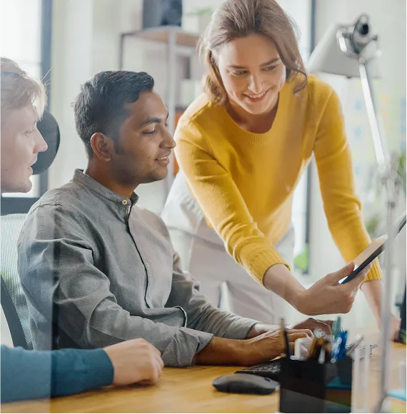 Group in a modern office working together on a screen; standing person points to a tablet, focus on teamwork, knowledge transfer and digital collaboration.