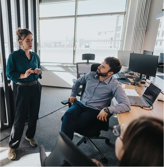 Collegial meeting in a modern office with daylight; standing person explains something to a colleague sitting opposite, in the background several workstations with monitors.