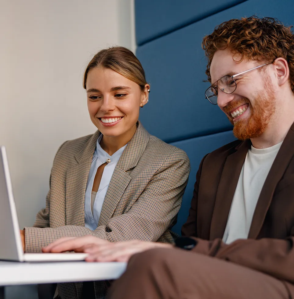 Two people laughing together while working on a laptop in a relaxed office environment; focus on team spirit, modern collaboration and positive corporate culture.
